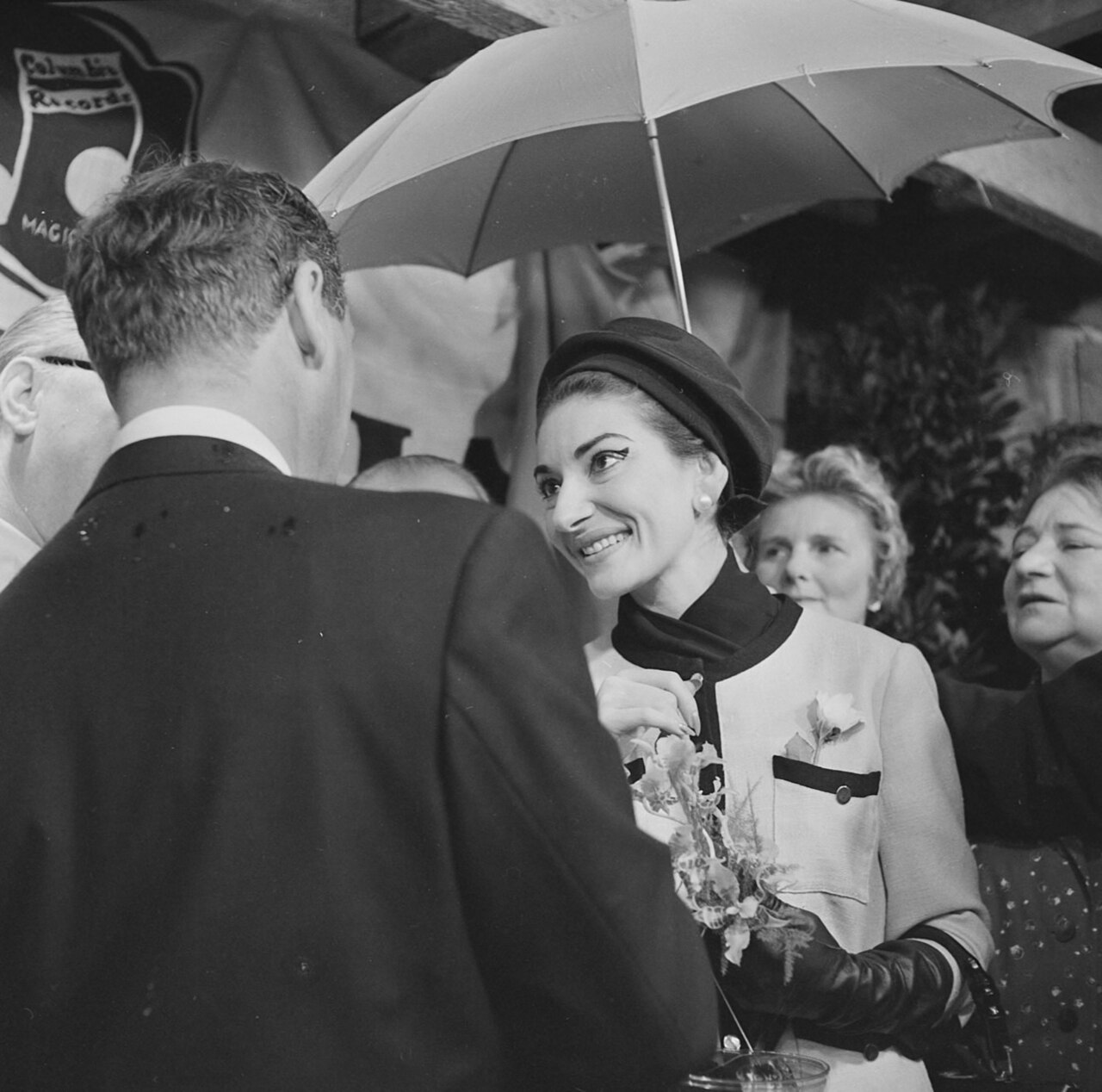 A woman in a stylish outfit and hat smiles whilst holding an umbrella and talking to a man in a suit. Like a scene from an opera, several people stand around them, some watching the conversation, in an outdoor setting.