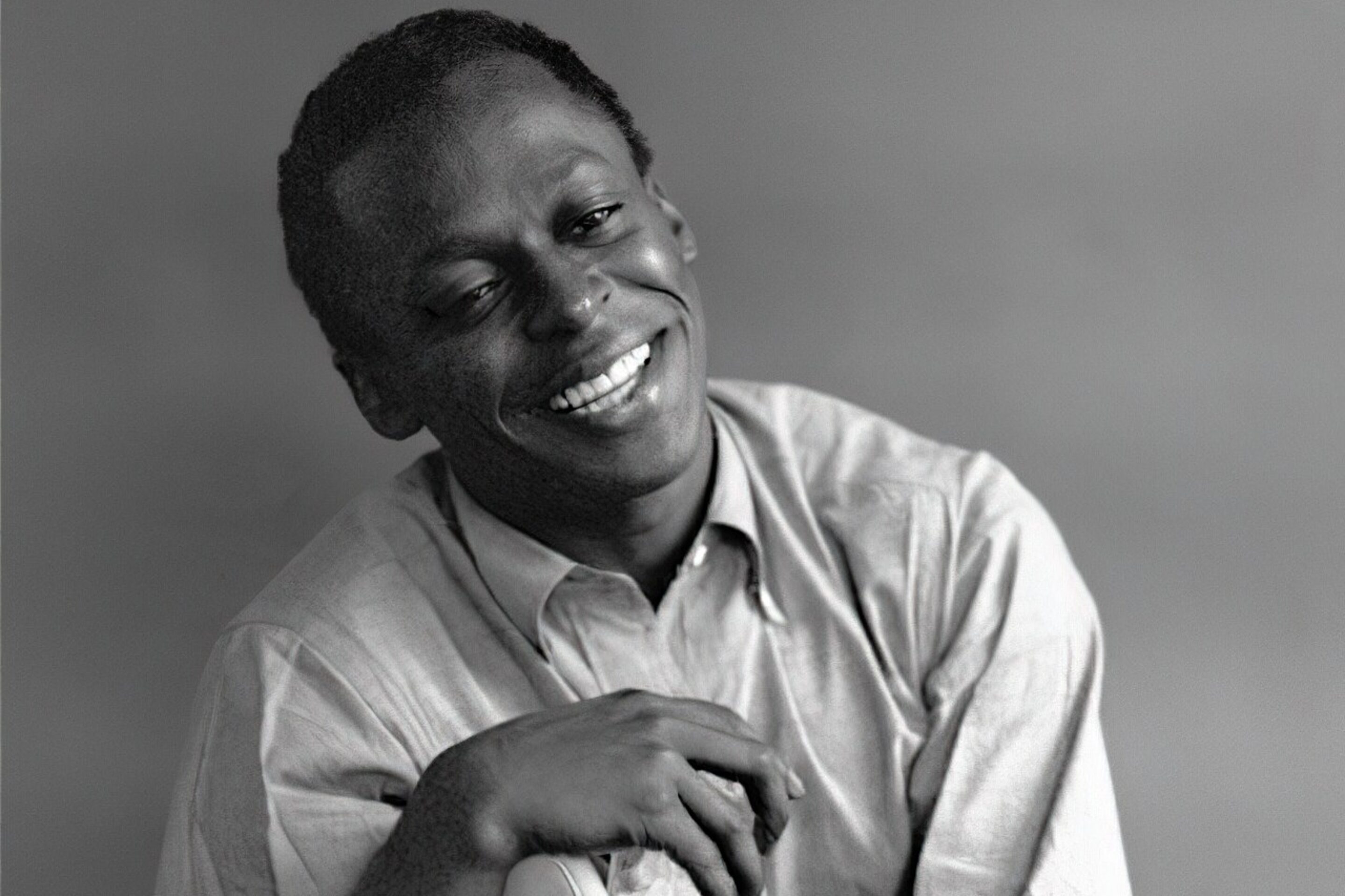 A smiling man in a button-up shirt leans casually on the back of a chair against a plain, softly blurred background, evoking classic portraits from history. The image is in black and white.