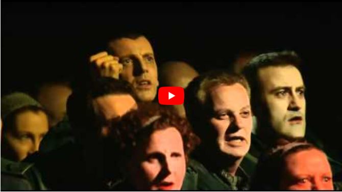 A group of people with sombre expressions and dramatic stage make-up sing together.