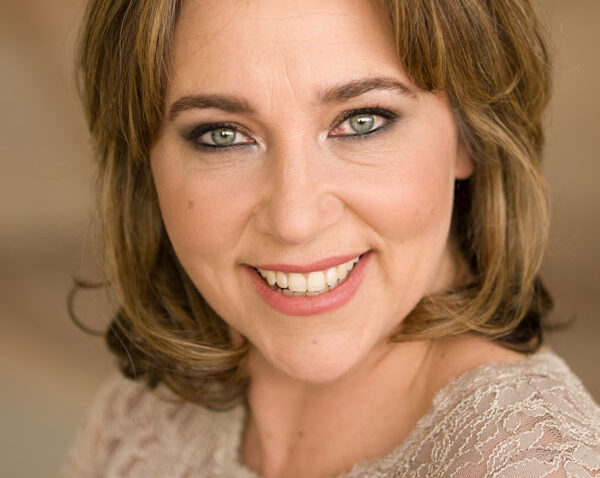 Christine Rice, a woman with shoulder-length light brown hair and green eyes, smiles warmly at the camera. She is wearing a lacy, cream-coloured top, and the background is softly blurred in neutral tones.