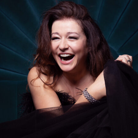 Carolyn Dobbin, with long brown hair, laughs joyfully whilst wrapped in sheer black fabric, sitting against a dark teal, radiating background. She wears a silver bracelet and has a radiant, expressive smile.