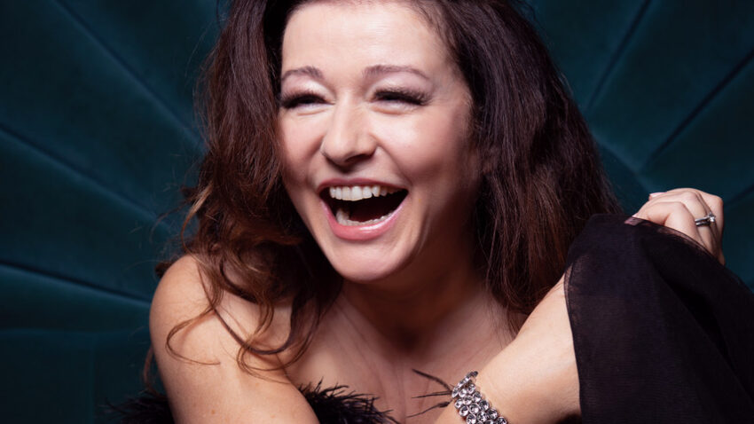 Carolyn Dobbin, with long brown hair, laughs joyfully whilst wrapped in sheer black fabric, sitting against a dark teal, radiating background. She wears a silver bracelet and has a radiant, expressive smile.