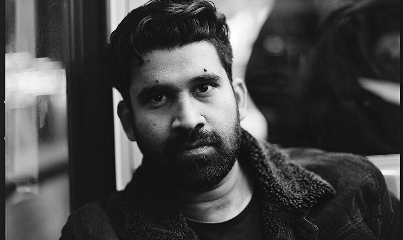 Black and white photo of Darwin Prakash, a bearded man with dark hair, wearing a jacket with a furry collar, sitting indoors and looking directly at the camera with a serious expression.