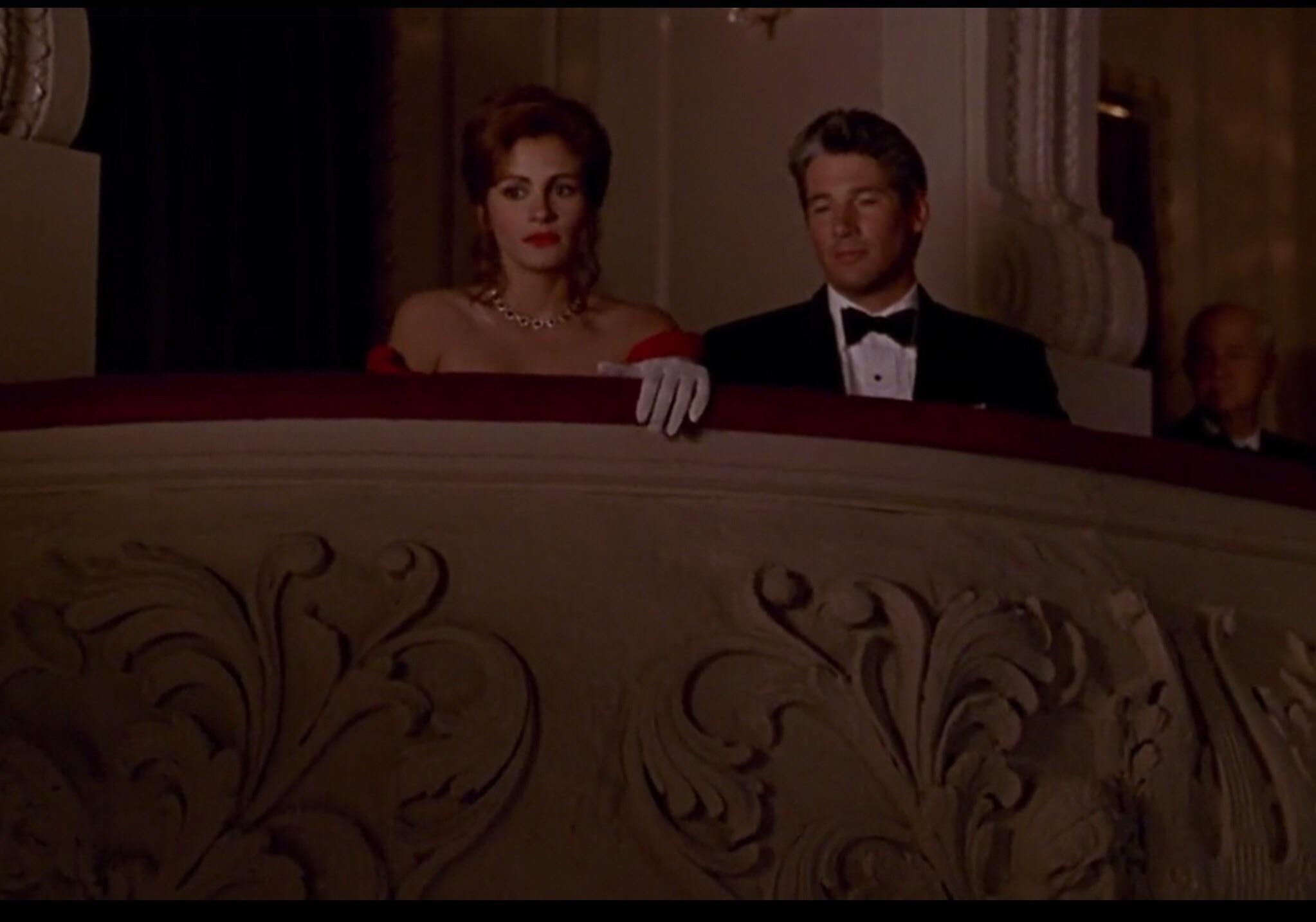 A woman in a red dress and white gloves sits beside a man in a black dinner jacket on a theatre balcony, both gazing at the stage during an introduction to La traviata, surrounded by elegant décor.