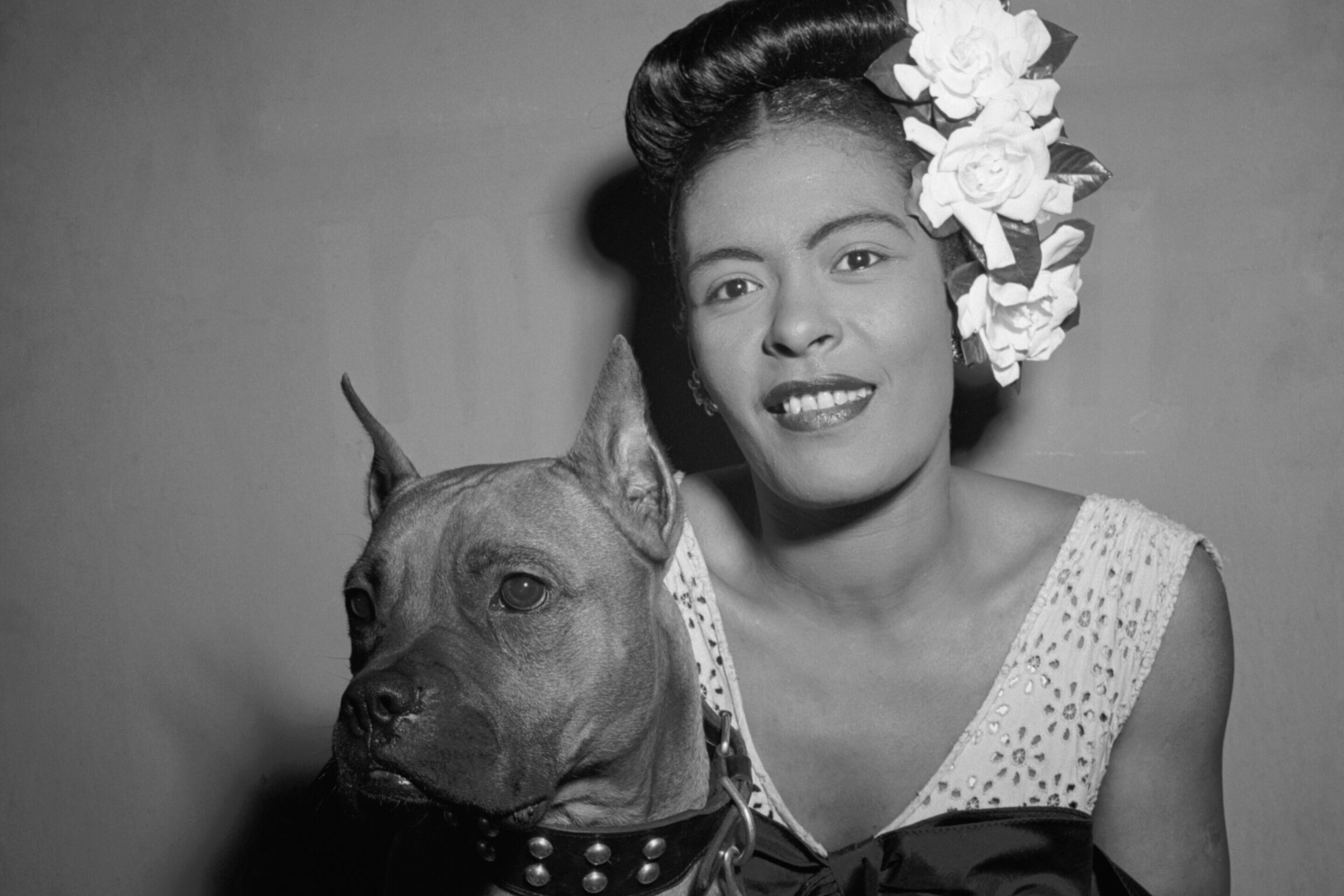 A woman wearing a floral hairpiece and elegant dress smiles whilst posing with her dog in a studded collar. The black-and-white photo evokes the timeless charm of Summertime, reminiscent of George Gershwin’s classic style.