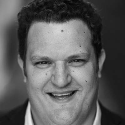 Black and white close-up portrait of a man with curly hair, smiling, wearing a dark jacket and light shirt, with a blurred background.