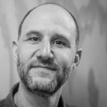 A black and white portrait of Adam Silverman, a middle-aged man with a trimmed beard and short hair, looking at the camera with a gentle expression. The background is blurred and softly textured.