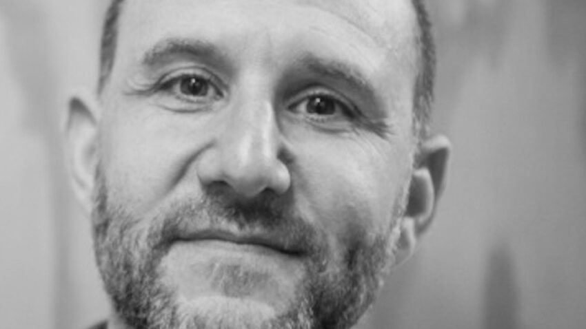 A black and white portrait of Adam Silverman, a middle-aged man with a trimmed beard and short hair, looking at the camera with a gentle expression. The background is blurred and softly textured.