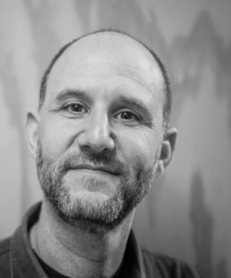 A black and white portrait of Adam Silverman, a middle-aged man with a trimmed beard and short hair, looking at the camera with a gentle expression. The background is blurred and softly textured.