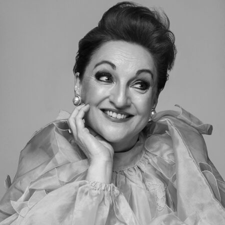 Ailish Tynan, with short, styled hair, smiles and gazes upwards. She wears a ruffled, sheer top, large pearl earrings, and has her hand resting on her cheek. The photo is in black and white.