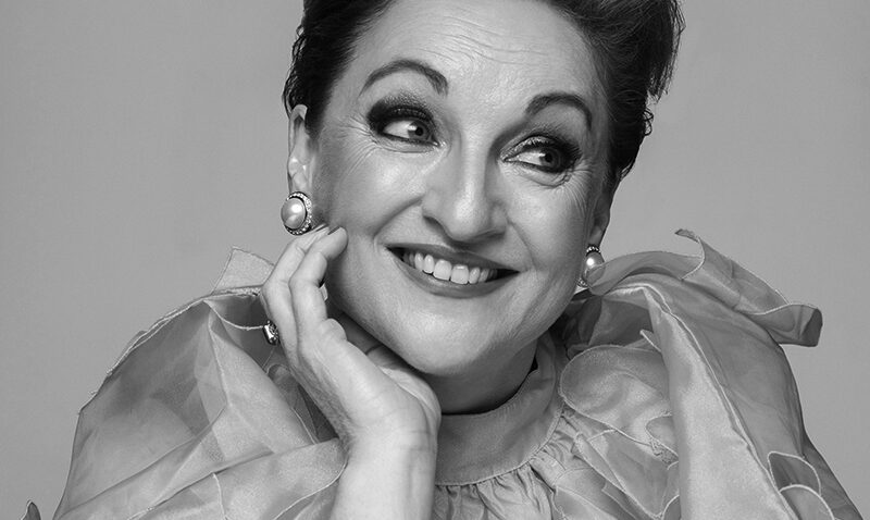 Ailish Tynan, with short, styled hair, smiles and gazes upwards. She wears a ruffled, sheer top, large pearl earrings, and has her hand resting on her cheek. The photo is in black and white.