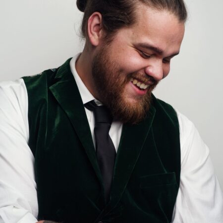 Alaric Green, a bearded man with a bun, wearing a white shirt, green velvet waistcoat, and black tie, smiles and looks down. He has a large black striped tattoo on his forearm against a plain light background.