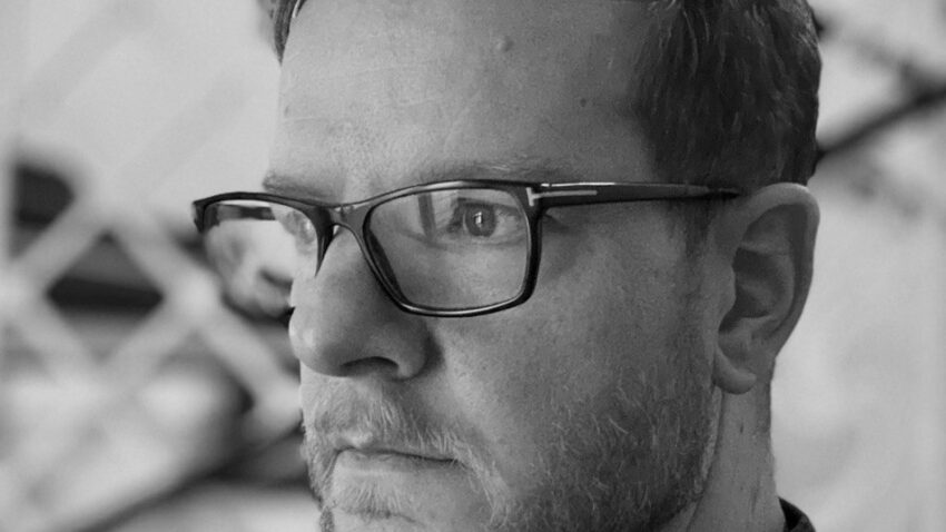 A man with short hair, a beard, and glasses looks to the side with a serious expression. He is wearing a checked shirt, and the background appears abstract and blurred. The photo is in black and white.
