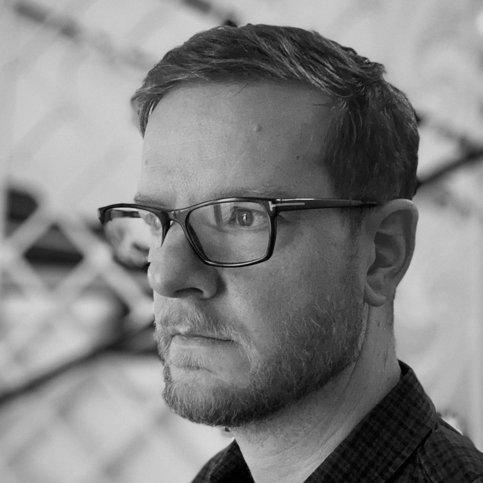 A man with short hair, a beard, and glasses looks to the side with a serious expression. He is wearing a checked shirt, and the background appears abstract and blurred. The photo is in black and white.
