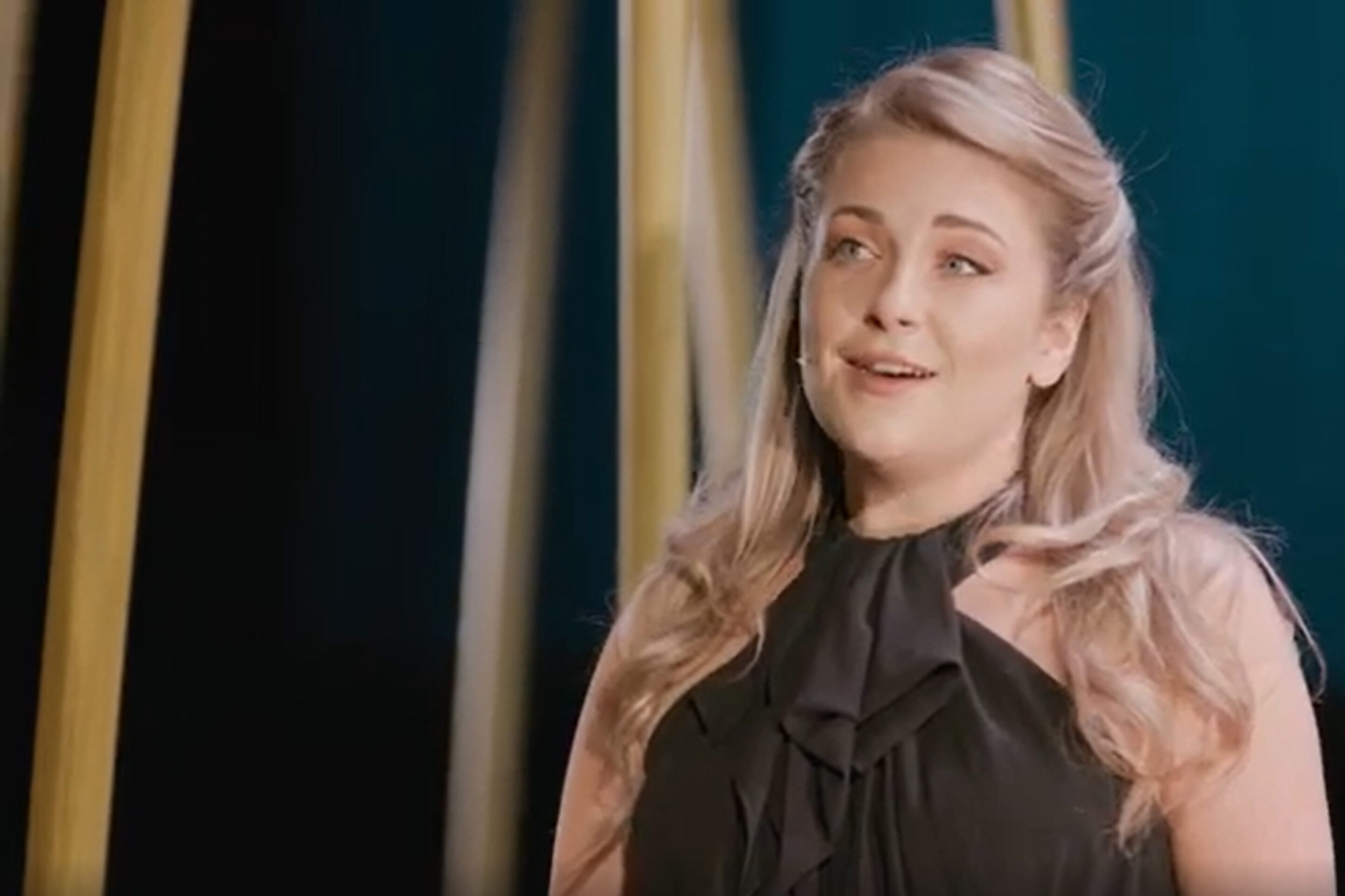 A woman with long blonde hair, wearing a sleeveless black top with a bow at the neck, stands on stage under soft lighting, appearing to sing in a performance.