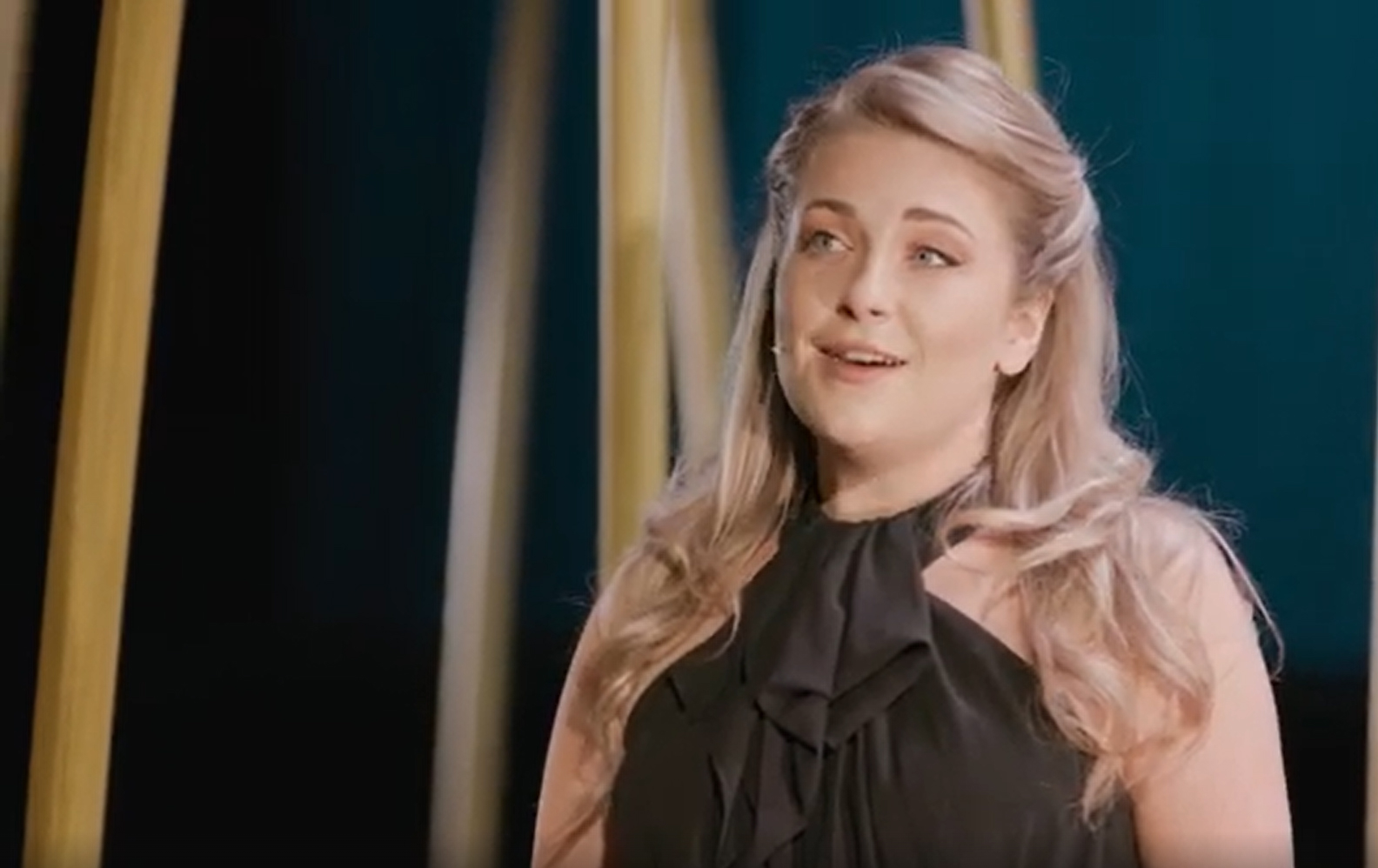 A woman with long blonde hair, wearing a sleeveless black top with a bow at the neck, stands on stage under soft lighting, appearing to sing in a performance.