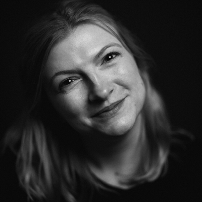 Black and white portrait of Amy Holyland with fair hair, smiling softly. She is facing the camera with a gentle expression, and the dark background makes her face the focal point of the image.