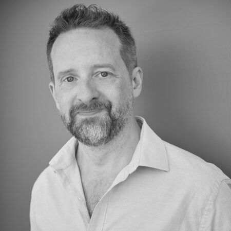 Andrew Foster-Williams, a man with short, dark hair and a beard, wearing a light-coloured, open-collar shirt, stands against a plain background, smiling gently. The photo is in black and white.