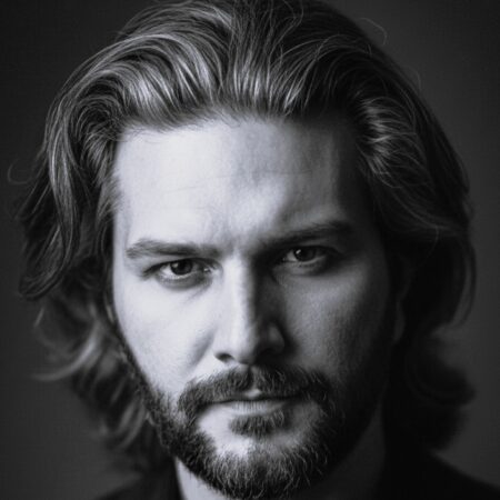 Black and white portrait of Andrew Manea, a man with medium-length wavy hair and a beard, looking directly at the camera with a serious expression. The background is dark and blurred.