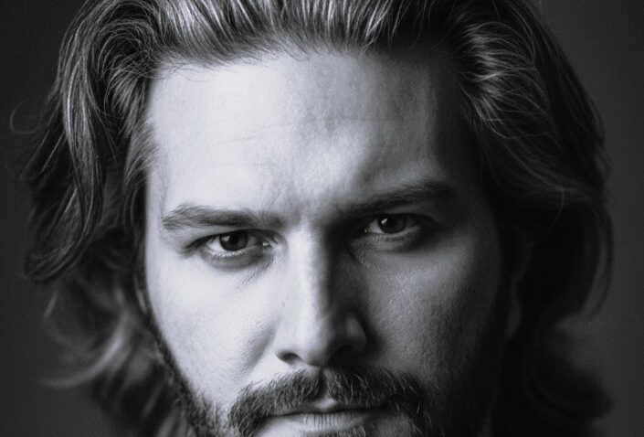 Black and white portrait of Andrew Manea, a man with medium-length wavy hair and a beard, looking directly at the camera with a serious expression. The background is dark and blurred.