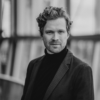 Black and white portrait of Andri Björn Róbertsson, a man with wavy hair and light stubble, wearing a checked blazer and polo neck, standing in front of a blurred modern architectural background.