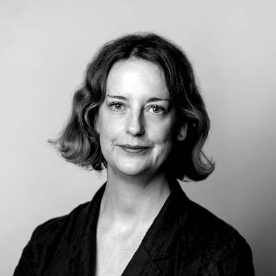 Black-and-white portrait of Annilese Miskimmon with wavy, shoulder-length hair, wearing a dark jacket. She gazes at the camera with a slight smile, set against a plain, light background.