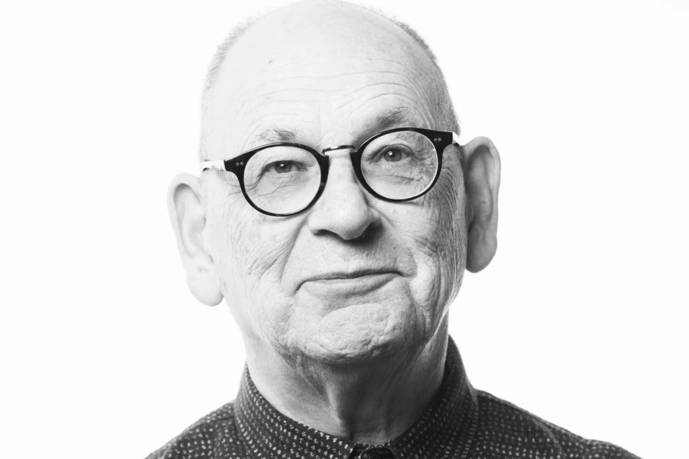 Black and white portrait of Antony McDonald, an older man with glasses, a bald head, and a patterned button-up shirt, looking directly at the camera with a slight smile against a plain white background.