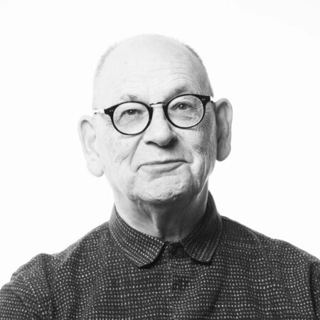 Black and white portrait of Antony McDonald, an older man with glasses, a bald head, and a patterned button-up shirt, looking directly at the camera with a slight smile against a plain white background.