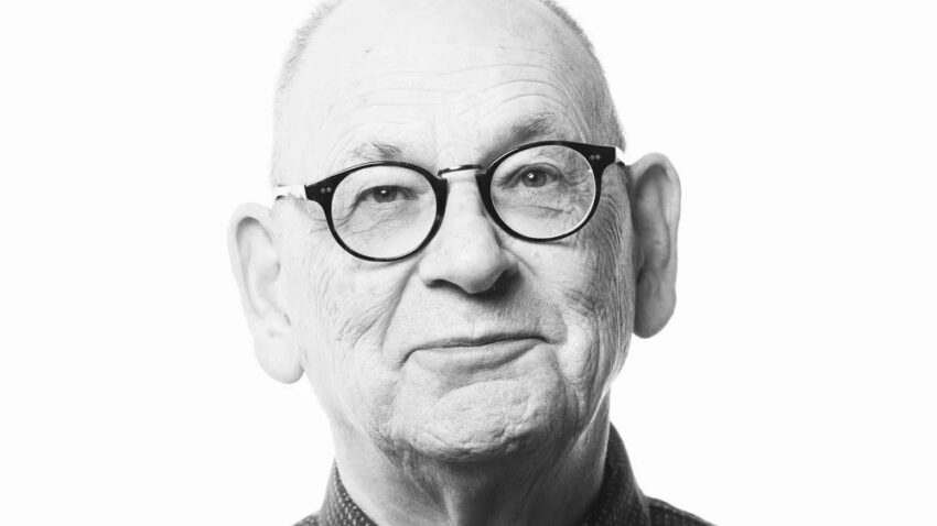 Black and white portrait of Antony McDonald, an older man with glasses, a bald head, and a patterned button-up shirt, looking directly at the camera with a slight smile against a plain white background.