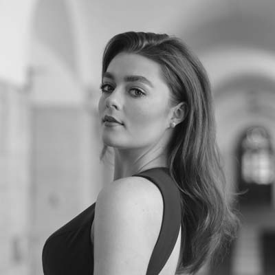 A woman with long, wavy hair wearing a sleeveless dress looks over her shoulder in a corridor with arched ceilings. The image is in black and white.