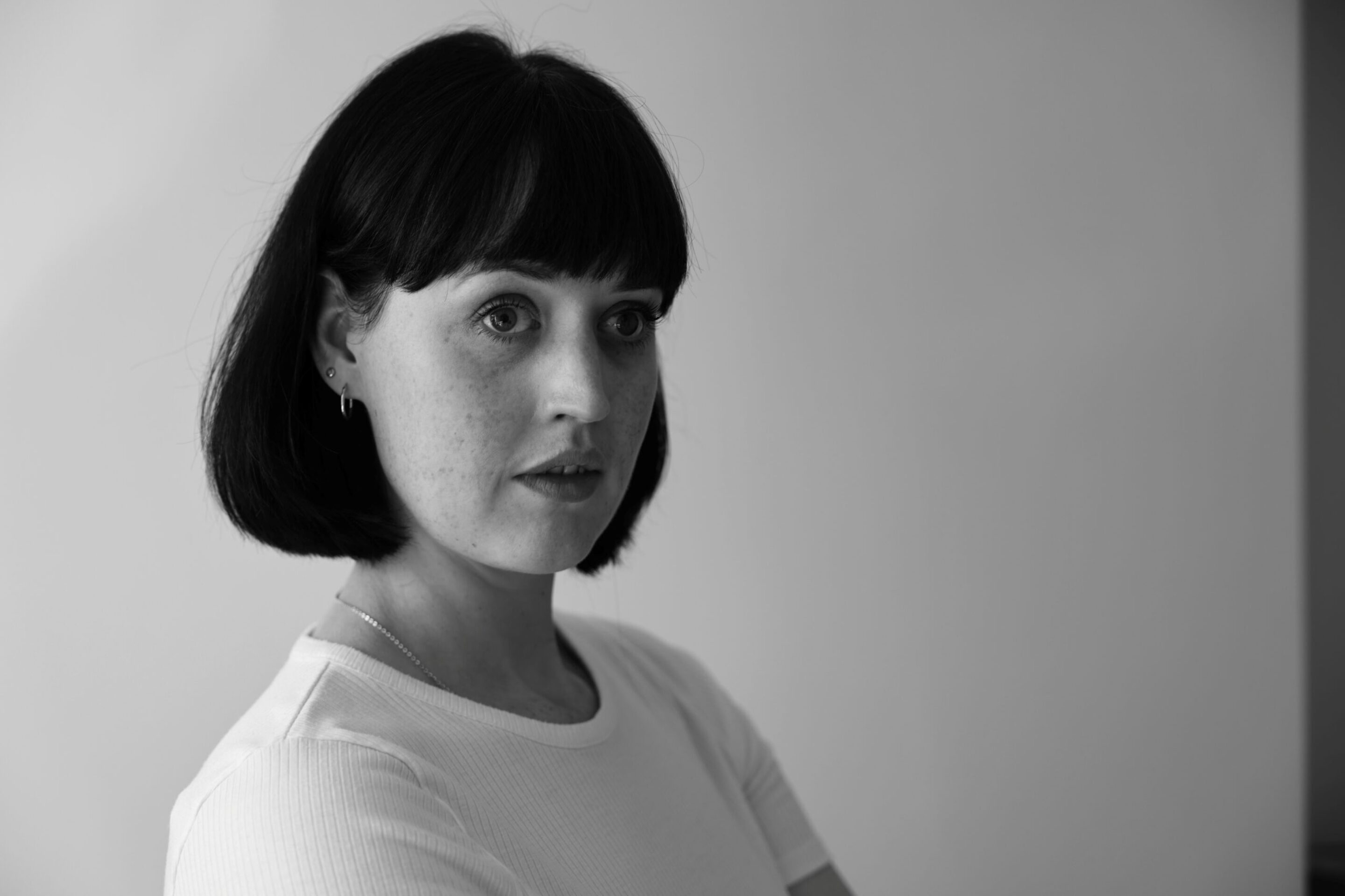 Bethan Langford, a woman with short dark hair and fringe, wearing a light-coloured shirt, looks off to the side with a thoughtful expression against a plain background. The image is in black and white.