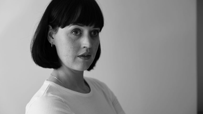 Bethan Langford, a woman with short dark hair and fringe, wearing a light-coloured shirt, looks off to the side with a thoughtful expression against a plain background. The image is in black and white.
