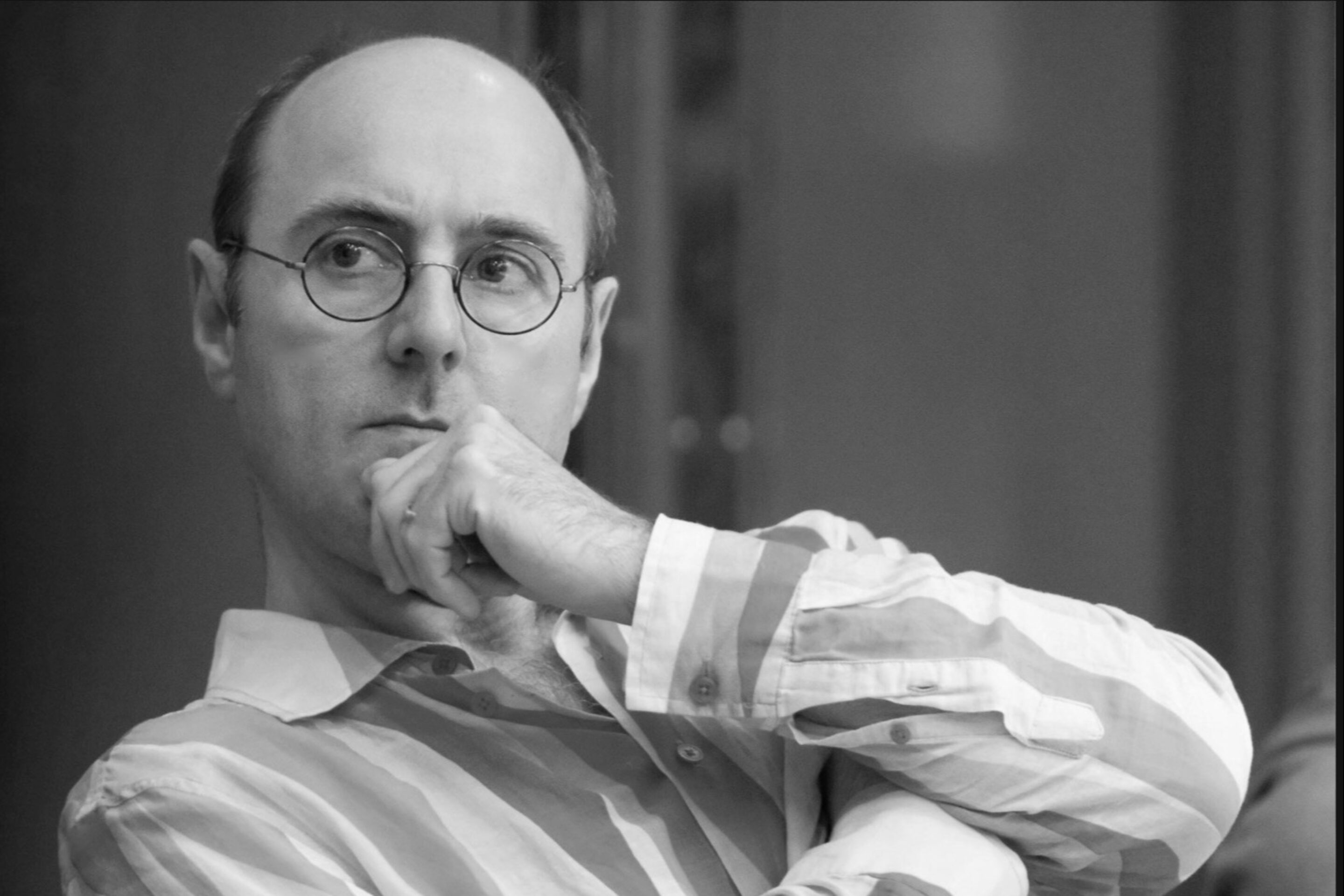 A man wearing glasses and a striped shirt, resembling Cal McCrystal, sits with his hand on his chin, looking thoughtfully to the side. The image is in black and white.
