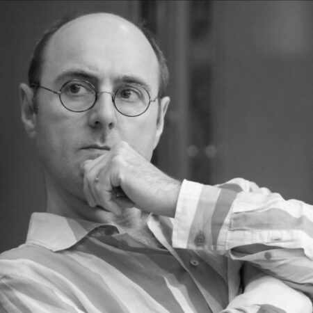 A man wearing glasses and a striped shirt, resembling Cal McCrystal, sits with his hand on his chin, looking thoughtfully to the side. The image is in black and white.
