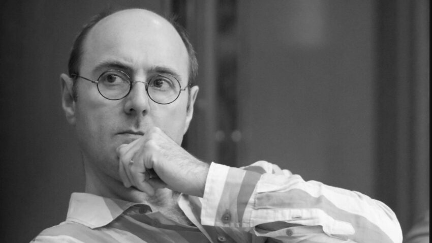 A man wearing glasses and a striped shirt, resembling Cal McCrystal, sits with his hand on his chin, looking thoughtfully to the side. The image is in black and white.