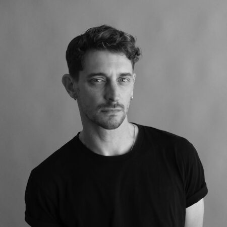 Black and white portrait of Cameron McMillan, a man with short curly hair wearing a dark t-shirt. He stands against a plain background, looking slightly to the side with a neutral expression.