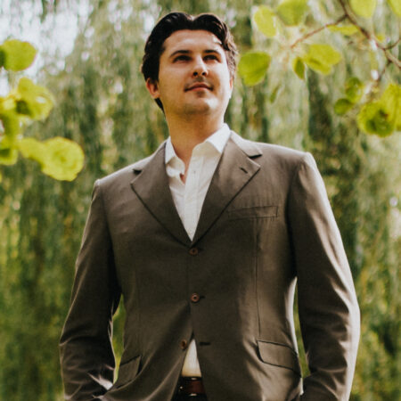 A man in a brown suit and white shirt stands outdoors among green leaves, looking upward with his hands in his pockets—Caspar Singh gazes thoughtfully as the background blurs with trees and foliage.