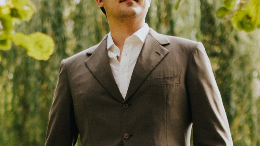 A man in a brown suit and white shirt stands outdoors among green leaves, looking upward with his hands in his pockets—Caspar Singh gazes thoughtfully as the background blurs with trees and foliage.