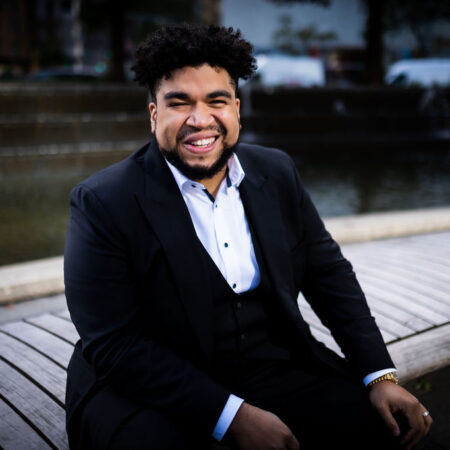 Chaz’men Williams-Ali, dressed in a dark suit, sits on a curved wooden bench by a fountain, smiling broadly at the camera. Behind him, trees and city buildings are slightly out of focus.