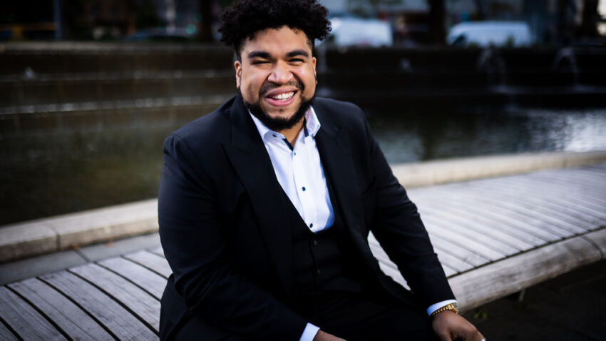 Chaz’men Williams-Ali, dressed in a dark suit, sits on a curved wooden bench by a fountain, smiling broadly at the camera. Behind him, trees and city buildings are slightly out of focus.