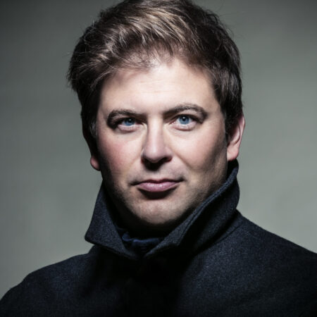 Craig Colclough, a man with light skin, blue eyes, and short brown hair, wears a dark coat with the collar turned up. He faces the camera with a neutral expression against a plain, muted background.