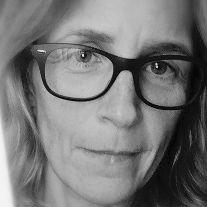Black and white close-up of a person with fair hair wearing large, dark-framed glasses, looking directly at the camera with a neutral expression.