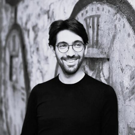 A smiling man with glasses and a beard, possibly Daniel Cohen, stands in front of a wall painted with large, vintage clock faces. The image is in black and white.