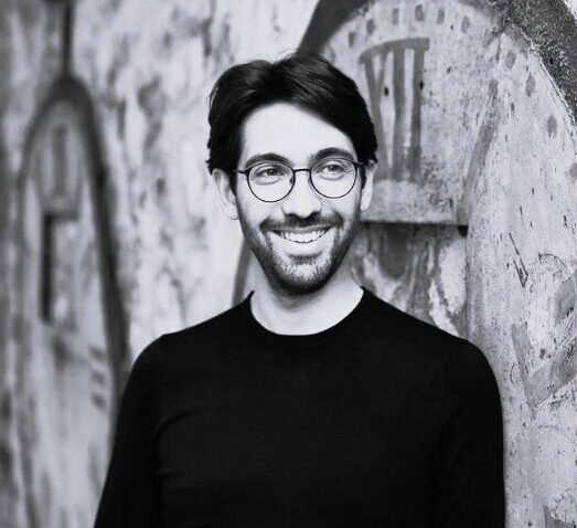 A smiling man with glasses and a beard, possibly Daniel Cohen, stands in front of a wall painted with large, vintage clock faces. The image is in black and white.