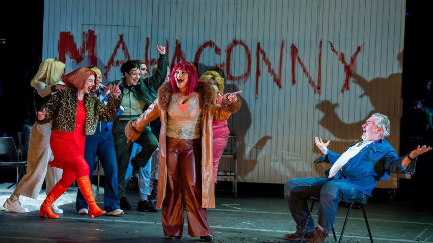 A group of actors perform energetically on stage; at centre, a woman in a brown outfit sings as the drama of Mahagonny unfolds. The word MAHAGONNY is smeared in red on the wall behind them, evoking Rise and Fall’s charged atmosphere.
