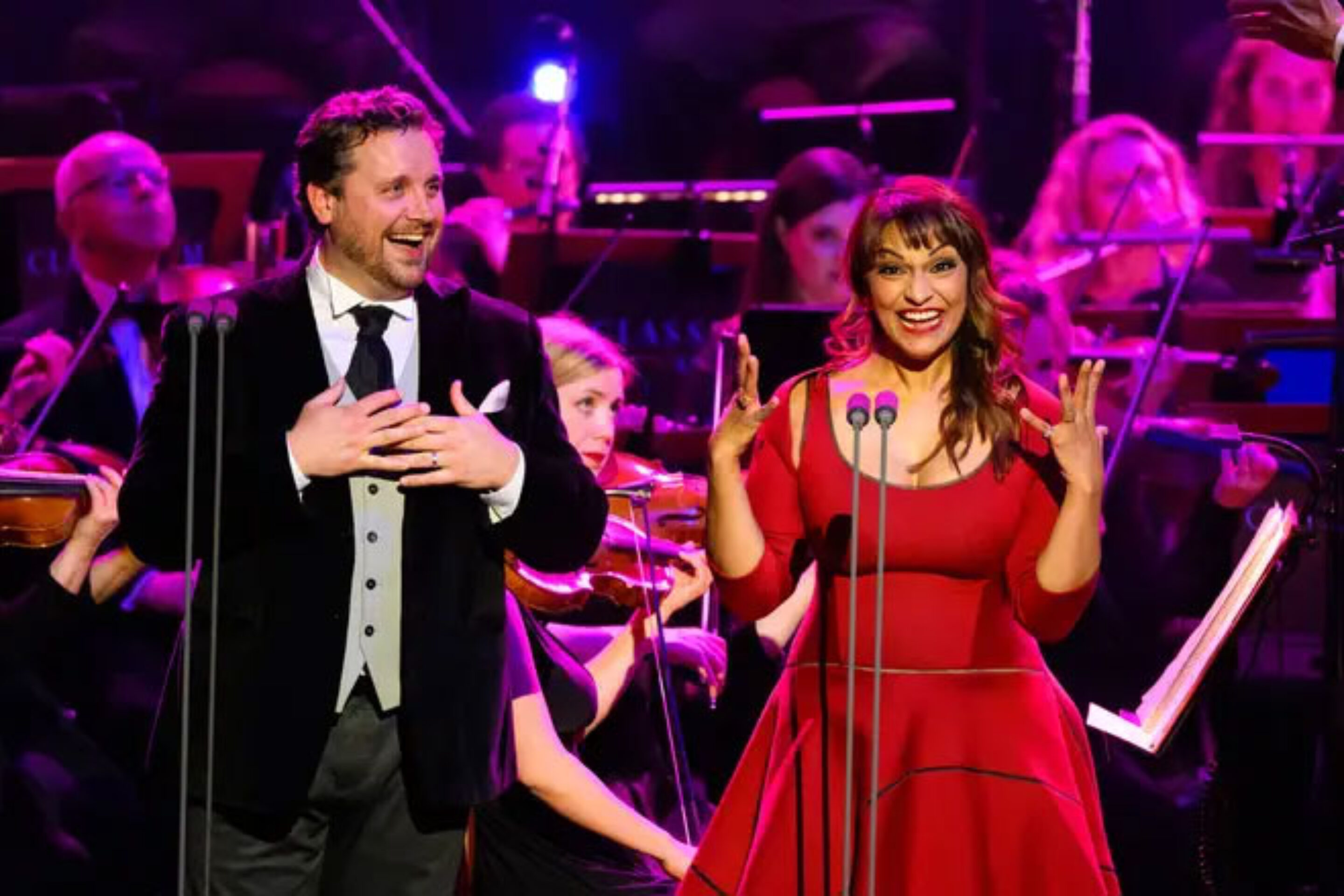 A man in a black dinner jacket and a woman in a red dress sing famous opera songs enthusiastically on stage, backed by an orchestra. Both have expressive gestures and joyful expressions under bright stage lighting.
