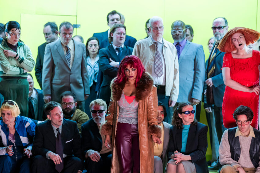 A woman with bright red hair, wearing a fur-trimmed coat and maroon trousers, stands confidently before a diverse group in eclectic attire, capturing the dramatic spirit of Mahagonny against a bold yellow background.