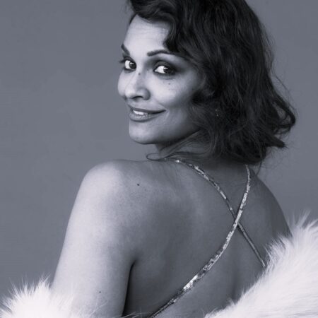 Soprano Danielle de Niese, with wavy hair, looks over her shoulder and smiles. She is wearing a strappy dress and a white faux fur stole. The photo is in black and white.
