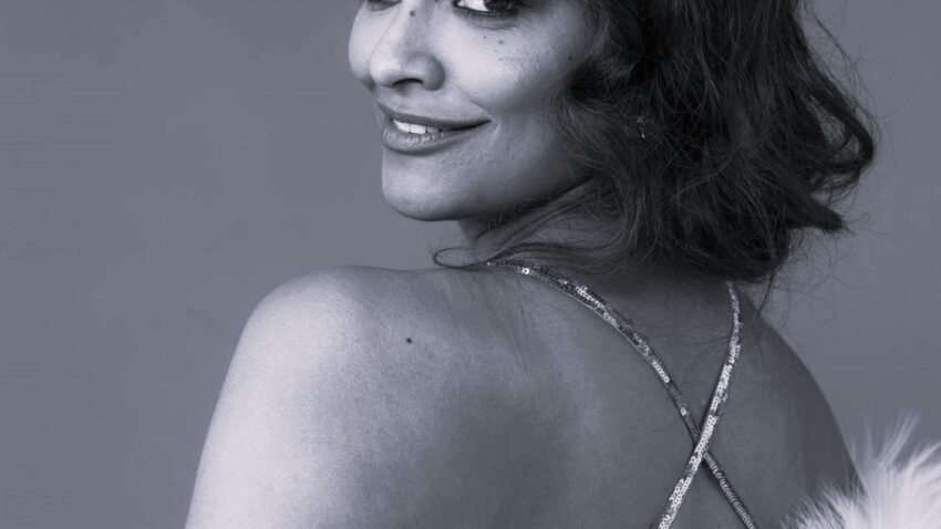 Soprano Danielle de Niese, with wavy hair, looks over her shoulder and smiles. She is wearing a strappy dress and a white faux fur stole. The photo is in black and white.