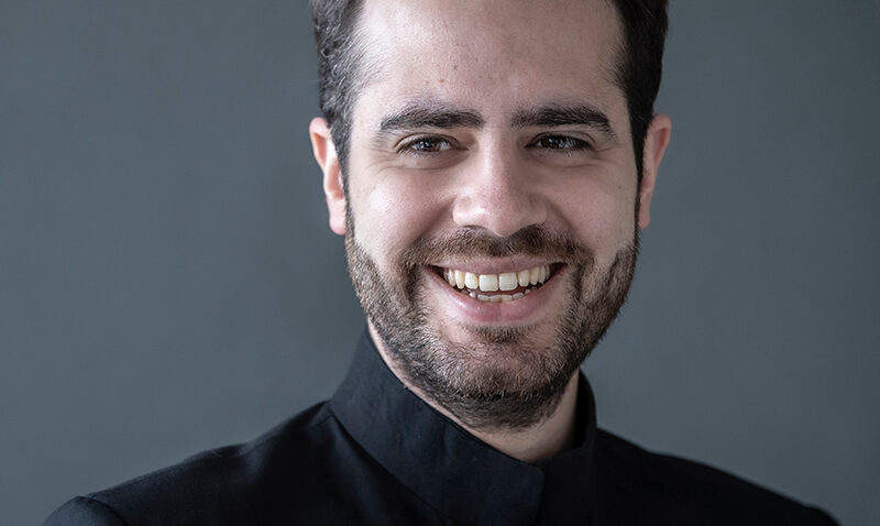 A man with short dark hair and a beard smiles at the camera. He is wearing a black jacket with a stand-up collar and is posed against a plain grey background.